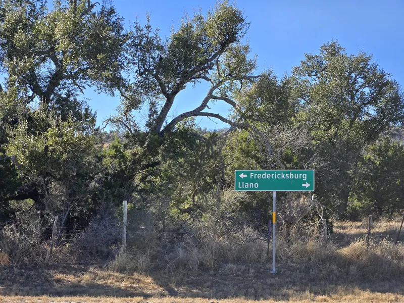 A road sign pointing toward Fredericksburg and Llano under live oaks.