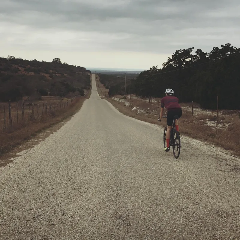 A rider on a long, straight Hill Country road under a gray sky.