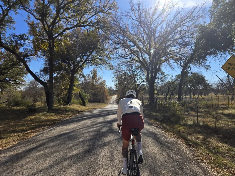 A rider in a white and maroon kit on a tree-lined road past a water crossing sign.