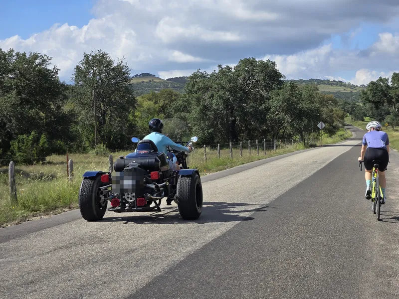 A three-wheeled motorcycle passing a rider on a Hill Country road.